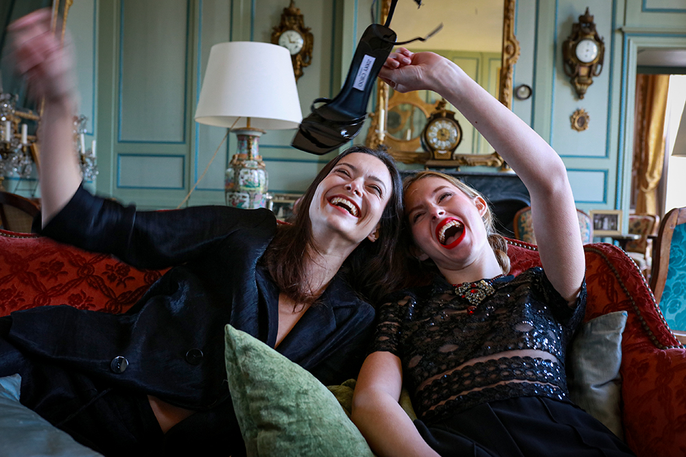 Deux femmes éclatent de rire dans un intérieur élégant, l’une tenant une chaussure à talon. Photographie © Thibaut Thouzery